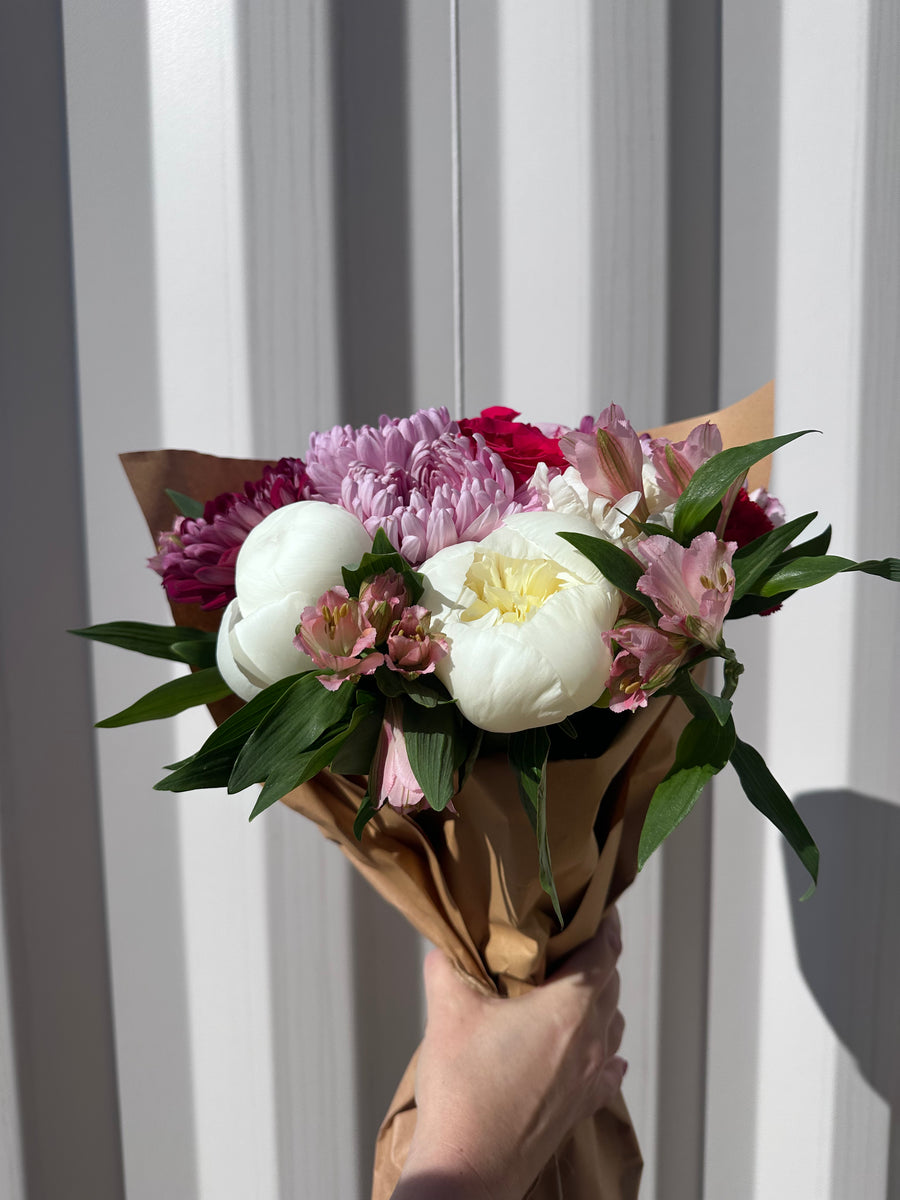 A wrapped Alegria bouquet of white peonies, pink roses and purple blooms held up against a white wall